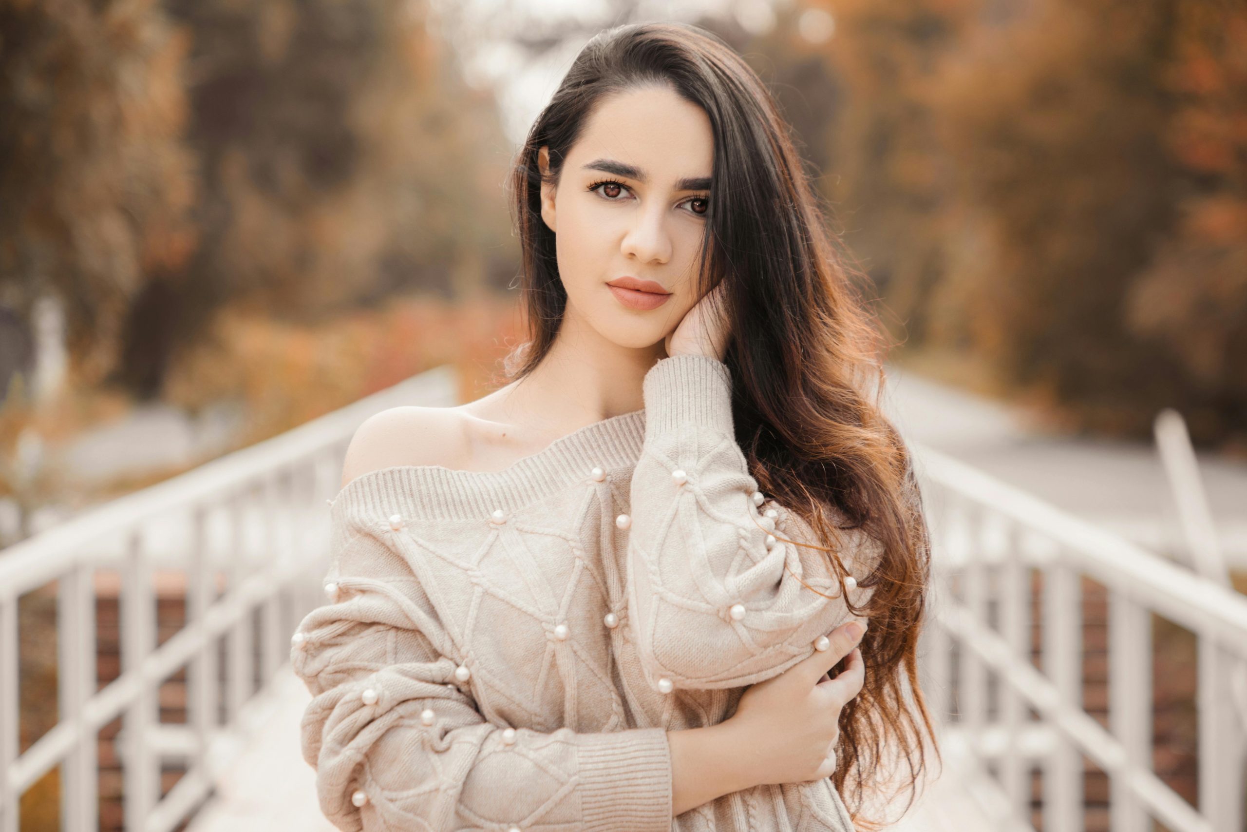 A young woman poses outdoors in a stylish sweater amidst fall foliage, exuding timeless beauty.